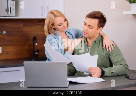 beautiful lovely young couple reading and analyzing bills sitting at table and using modern laptop. Young couple in casual discussing home economics Stock Photo