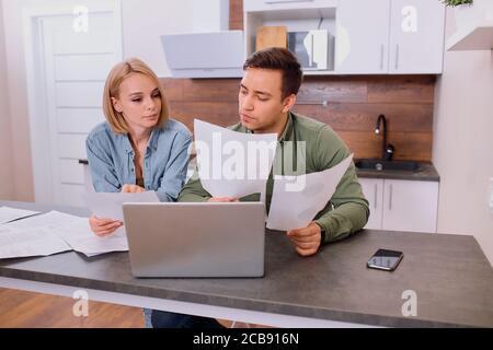 thoughtful couple solving financial problems in family, using laptop, holding paper documents, discussing costs for past month Stock Photo