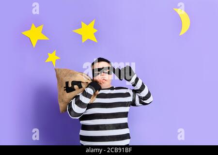criminal man in mask holding gun weapon near his temple , looking up, planning his crime over isolated blue background with moon and stars. Stock Photo