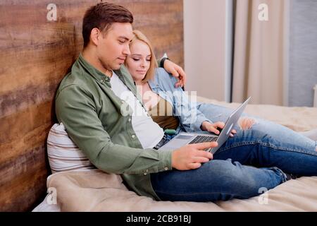 young caucasian couple lying on bed and watching film, in casual wear, looking at screen of modern laptop, spend free time together at home Stock Photo