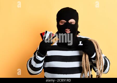 prisoner in mask and rope on his houlder showing credit and shopping cards. isolated yellow background. studio shot. Stock Photo