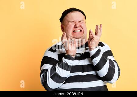 nervous burglar with raised hands wearing striped clothes in panic, shouting , screaming, isolated yellow background.negative emotion Stock Photo