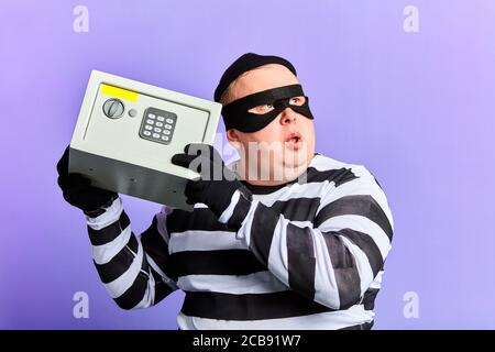 criminal thief or burglar man in mask holding a safe in hand for break opening it. close up portrait, isolated blue background. studio shot. Stock Photo