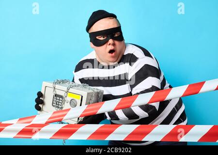 surprised scared burglar trying to hide safe from policemen. isolated blue background, studio shot Stock Photo