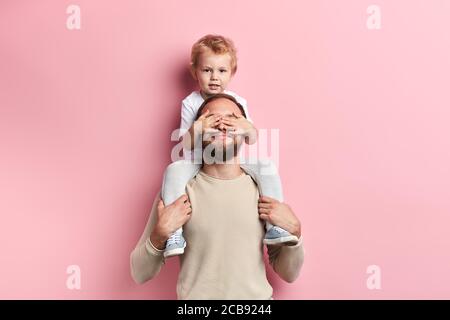 little boy closing his dad's eyes with palms, close up portrait, isolated pink background, studio shot Stock Photo