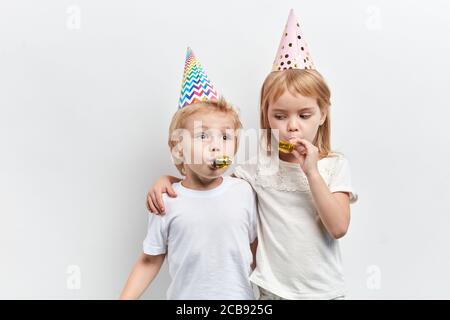 two cute children using party whistles, wearing party hats on a white background, happy childhood, close up portrait,holiday Stock Photo