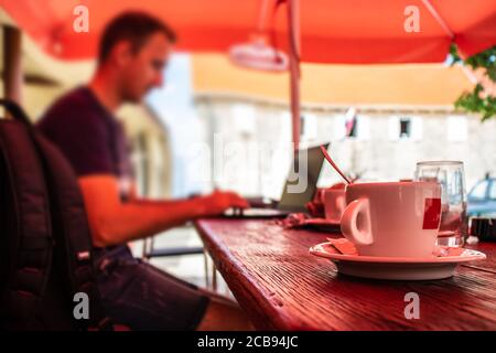 Closeup of a cup of coffee at a dalmatian coffee shop, blurred man sitting on a laptop working, concept of software developers working while travellin Stock Photo