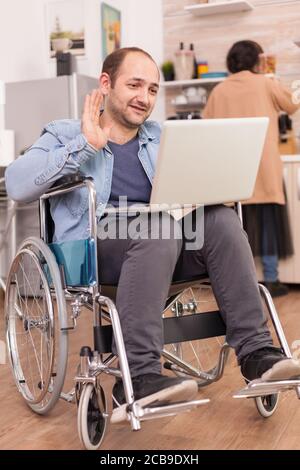 Disabled entrepreneur in wheelchair waving during a video call on laptop while wife is cooking lunch. Disabled paralyzed handicapped man with walking disability integrating after an accident. Stock Photo