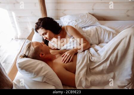 A young man and woman are basking in bed together. They are lying under white sheets. A young man and woman are basking in bed together. They lie under white sheet. Look at each other with tenderness. Stock Photo