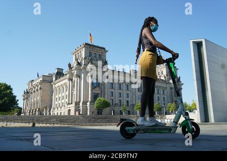 Berlin, Germany. 12th Aug, 2020. A woman with a mouth guard drives past the Reichstag building on an electric scooter. Today it is supposed to be up to 35 degrees. Credit: Jörg Carstensen/dpa/Alamy Live News Stock Photo