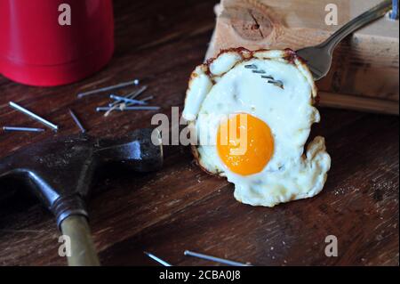 Closeup of fried egg hanging from a fork among carpenter tools. Hammer, nails and breakfast on wooden table. Stock Photo