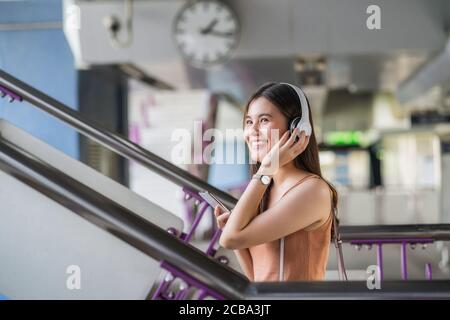 Young Asian woman passenger wearing headphone and using smart mobile phone to listening music and walking up the stairs in subway station when traveli Stock Photo