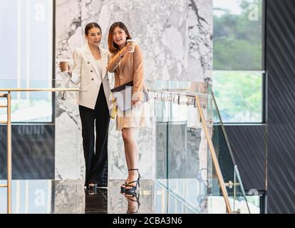 Two asian businesswomen walking and talking during coffee break in modern office or coworking space, coffee break, relaxing and talking after working Stock Photo