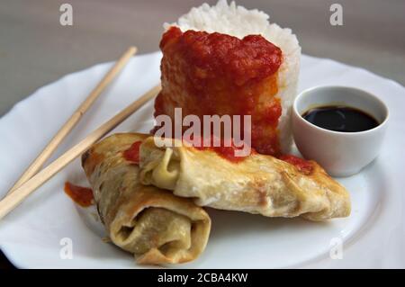 A plate with boiled rice with fried tomato, two spring rolls next to a bowl with soy sauce and some chopsticks Stock Photo
