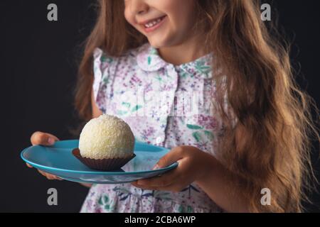 Beautiful little girl surprised from a tasty big chocolate coconut candy as a ball. Kid ready to eat cake. Stock Photo