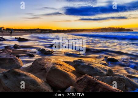 Sunrise at famous Bondi beach of Sydney on Pacific coast facing sandy beach and rocks in warm sun light.