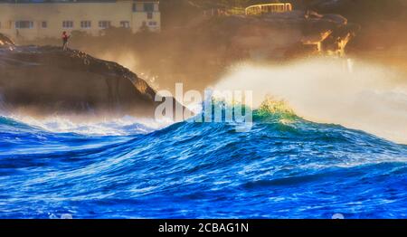 Bronte beach cliffs and strong waves of Pacific ocean in sun light around lonely fisher on a sandstone rock . Stock Photo