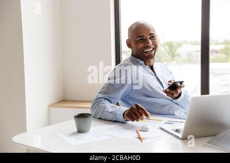 Photo of laughing african american man using cellphone while working with laptop at table in living room Stock Photo