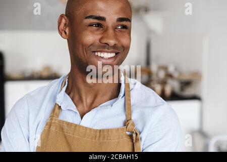 Photo of cheerful african american man in apron smiling and looking at camera at kitchen Stock Photo