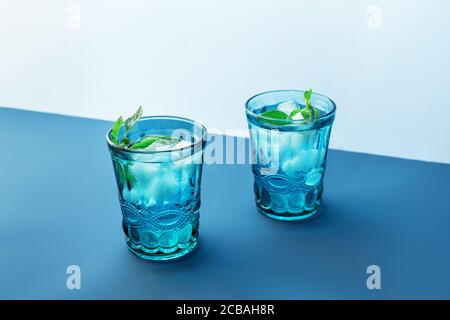 Two beautiful vintage blue glasses with cold drink and ice cubes, with fresh green mint leaves on deep blue and light blue background. Backdrop for your design. Stock Photo