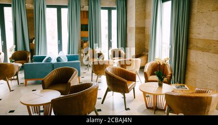 A large hall with exits to the balcony with round tables and flowers and brown armchairs made of genuine leather and blue sofas in the center. Stock Photo