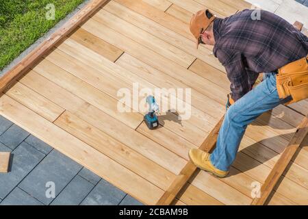 Caucasian Men in His 40s Building Wooden Deck on His Backyard Attaching Wood Board To Each Other Using Cordless Drivers and Screws. Construction Indus Stock Photo