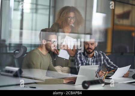 Helping coworkers. Three young happy colleagues using laptop and discussing work or sharing fresh ideas while working together in the modern office. Stock Photo