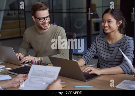 Working together. Young happy female and male colleagues, happy coworkers working on laptops and communicating while sitting in the modern coworking Stock Photo