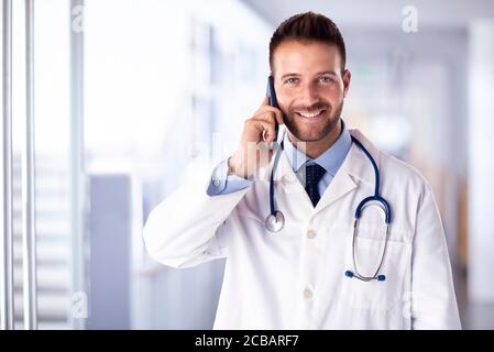 Shot of happy male doctor using phone and talking with somebody while standing on the hospital’s foyer. Stock Photo
