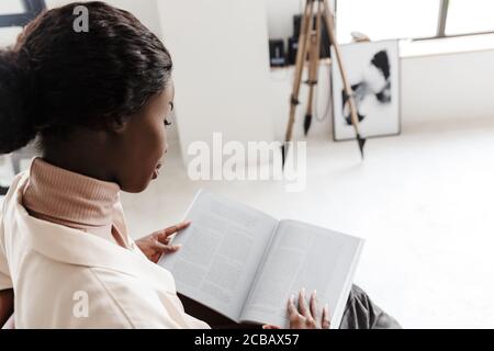 Side view photo of young african woman sitting on sofa indoors at home while reading magazine or book Stock Photo