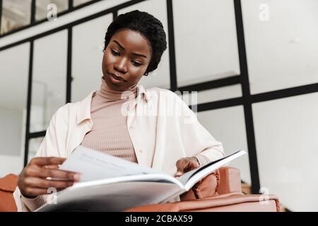 Photo of pretty concentrated young african woman sitting on sofa indoors at home while reading magazine or book Stock Photo