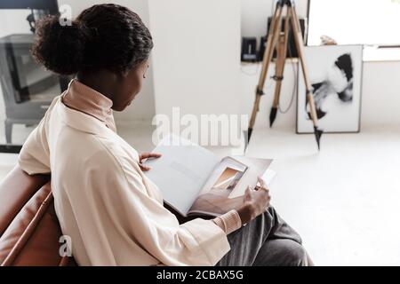 Back view picture of young african woman sitting on sofa indoors at home while reading magazine or book Stock Photo