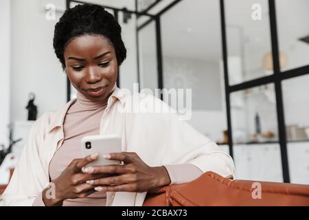 Image of amazing concentrated young african woman sitting on sofa indoors at home while using mobile phone Stock Photo