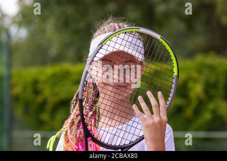 An aspiring smiling girl tennis player looks through a tennis racket. Stock Photo