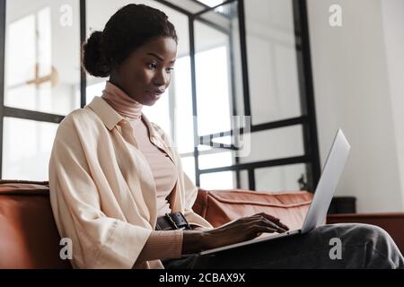 Photo of young concentrated woman sitting on sofa indoors at home while working with laptop computer Stock Photo