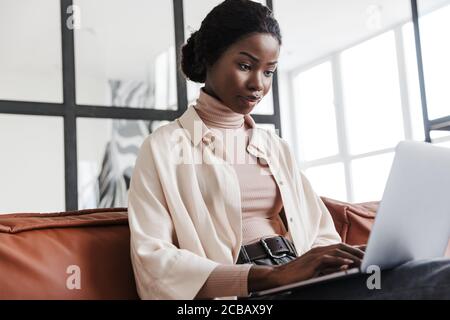 Photo of young serious woman sitting on sofa indoors at home while working with laptop computer Stock Photo