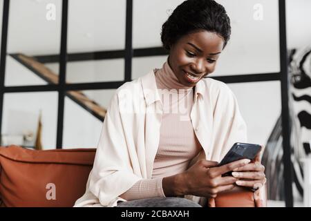 Image of amazing happy positive young african woman sitting on sofa indoors at home while using mobile phone Stock Photo