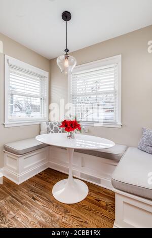 A small breakfast book in a residential home. The area has bench seating, cushions, and pillows with a small white oval table with flowers on top. Stock Photo
