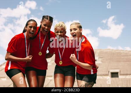 Female football team celebrating the victory at stadium. Woman soccer players with medals shouting in joy after winning the championship. Stock Photo