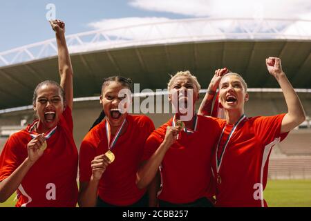 Woman soccer players with medals shouting in joy after winning the championship. Female football team celebrating the victory at stadium. Stock Photo