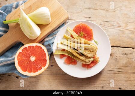 Sauteed fennel vegetable with grapefruit slices, vegetarian appetizer on a white plate, ingredients and a blue kitchen towel on rustic wood, copy spac Stock Photo