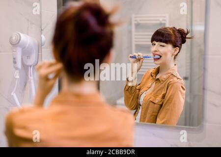Horizontal shot of young red haired smiling woman having fun in bathroom, winking to the mirror, brushing teeth and enjoying morning hygiene