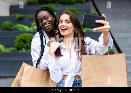 Two young women standing outdoors with shopping bags and make selfie on smartphone Stock Photo