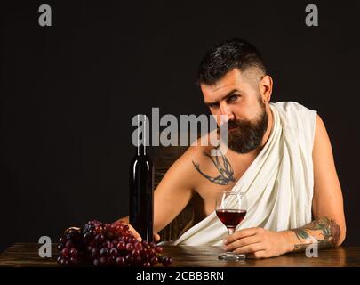 God of wine with tricky face wearing white cloth sits by wine bottle and grapes. Winemaking and degustation concept. Man with beard holds glass of alcohol on brown background. Sommelier tasting drink Stock Photo