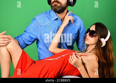 Pleasure, music and fun creative lifestyle concept. Music fans with serious faces enjoy music. Mans face with beard holds girl lying on green background. Couple in love wears headphones and sunglasses Stock Photo