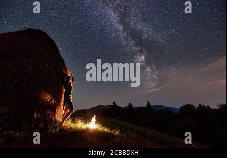 Silhouettes of girls near a haystack and a bonfire against the backdrop of a valley of hills, under incredibly beautiful starry sky on which the Milky Way is visible at night Stock Photo