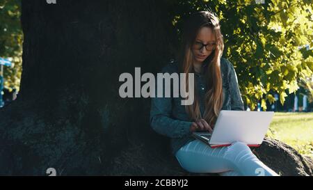 Young caucasian woman working on the laptop sitting near the tree in the park. High quality photo Stock Photo