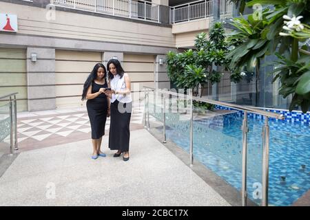 Two Indian woman having a discussion as they look into their phone in an urban corporate setting. Stock Photo