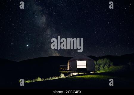 Beautiful gloving Yurt nomadic house at camping in the mountain valley under night sky with stars and Milky Way Stock Photo