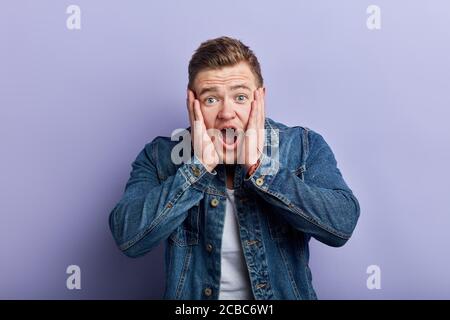 Stressful terrified young bearded man with wide open mouth covers mouth with palms , being shocked noticing huge spider on floor, expresses negative e Stock Photo
